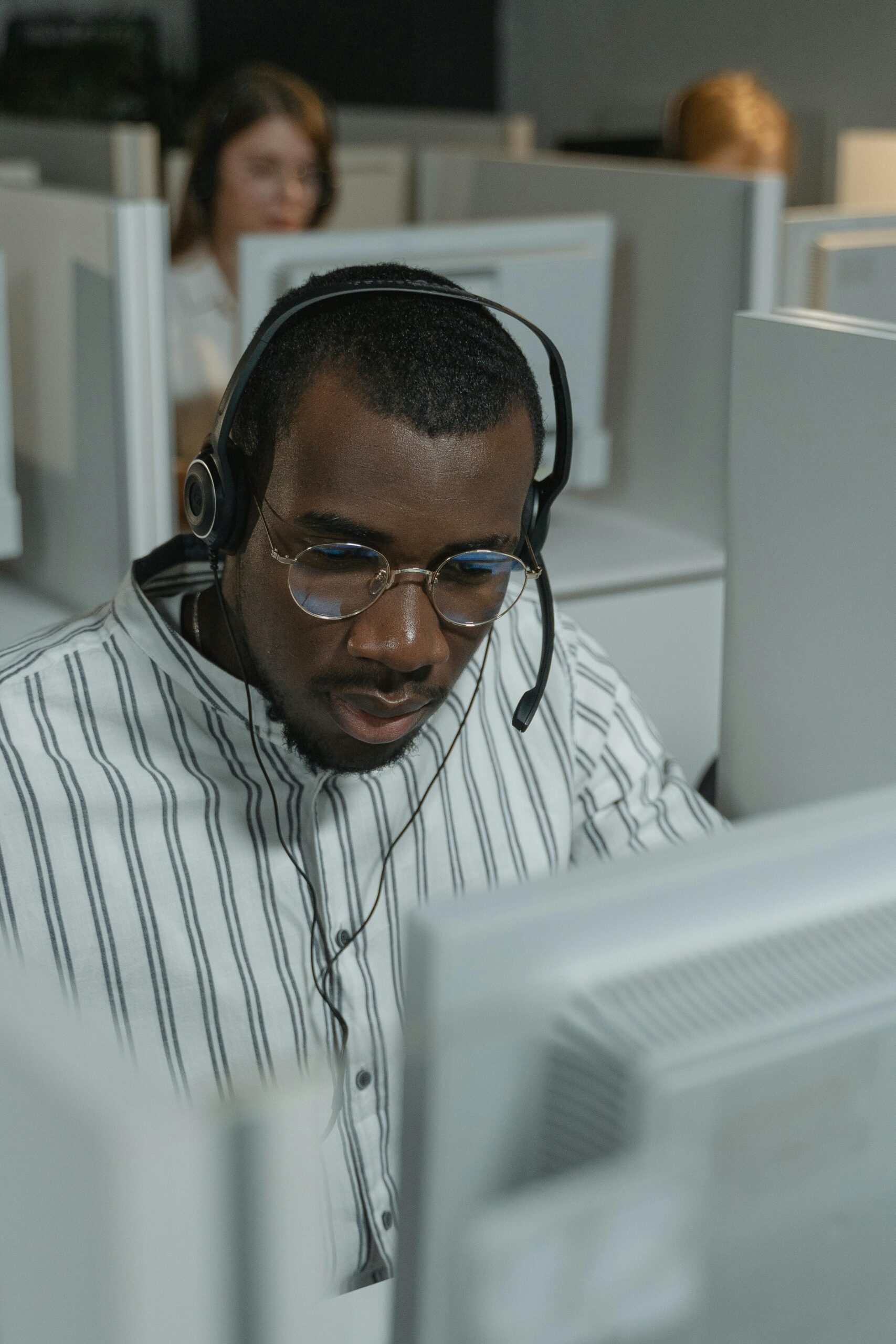 Customer support representative wearing glasses and headset in a busy call center setting.